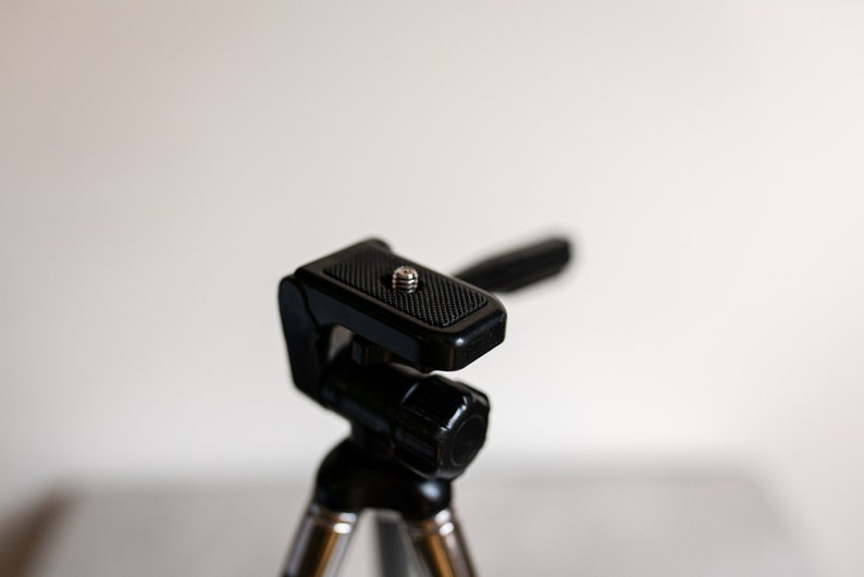 Puede incluir: Un tr&iacute;pode negro con una cabeza texturizada y ajustable. La cabeza tiene un tornillo para sujetar una c&aacute;mara u otro dispositivo.