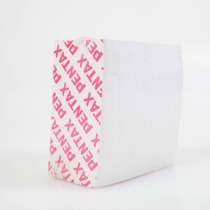 May include: A white rectangular product, possibly a camera accessory, wrapped in clear plastic. The side of the product features the word "PENTAX" in pink, repeated diagonally. The background is plain white.