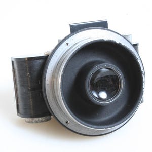 May include: A vintage black and silver camera with a round lens and a black body. The camera has a silver ring around the lens and a black strap attached to the top.