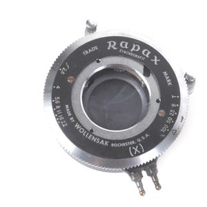 May include: A circular silver and black Rapax Synchromatic camera shutter mechanism by Wollensak, Rochester, U.S.A. The device features a central aperture, a knurled outer ring, and markings for f-stops from 2.8 to 22 and shutter speeds from 100 to T.