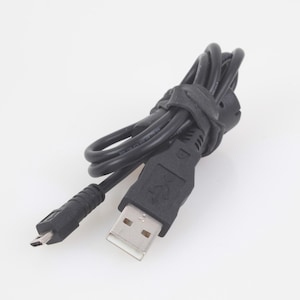 Puede incluir: Un cable USB negro con un conector micro USB y un conector USB estándar. El cable está enrollado y asegurado con una correa negra. Los conectores son negros con contactos metálicos plateados.
