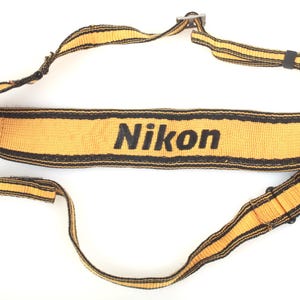 May include: Yellow and black Nikon camera strap. The strap has the word "Nikon" printed in black. The strap is made of a woven material and has black stripes along the edges. The strap is designed to be worn around the neck.
