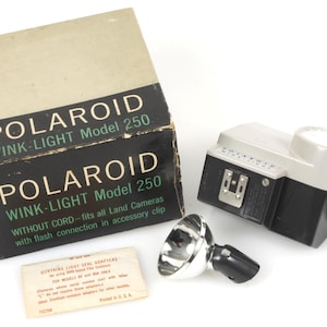 May include: A black and silver Polaroid Wink-Light Model 250 flash unit with a white reflector. The flash unit is in its original box, which is white with black lettering. The box reads "Polaroid Wink-Light Model 250".