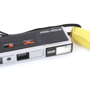 以下が含まれることがあります： 内蔵フラッシュと黄色のケースが付いた、黒と銀のミノルタオートパック450Eカメラです。