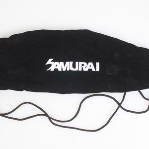 May include: Black drawstring bag with the word "SAMURAI" in white lettering. The bag is made of a soft, velvety material and has a drawstring closure with a black bead. The drawstring is black and extends from the bag.