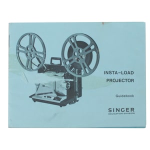 Könnte beinhalten: Ein blaues Handbuch für einen Insta-Load-Projektor. Das Cover zeigt einen schwarzen und silbernen Projektor mit zwei Filmrollen. Der Text "Insta-Load Projector" und "Guidebook" ist in Schwarz gedruckt. Das Logo der Singer Education Division ist unten in Schwarz gedruckt.