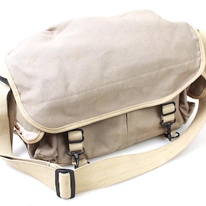 Puede incluir: Un bolso de mensajero de lona beige con una correa larga ajustable y múltiples hebillas. El bolso tiene un cierre de solapa y un bolsillo con cremallera en la parte delantera.