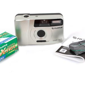 May include: A silver Fujifilm Smart Shot Plus camera with a black lens and flash, alongside a green and blue Fujifilm Superia X-TRA 400 film box and a manual. A black camera strap is also included.