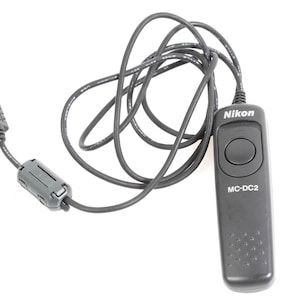 May include: A black Nikon MC-DC2 remote shutter release with a coiled black cable and a connector. The remote has a large round button and the Nikon logo. The model number MC-DC2 is printed on the remote.