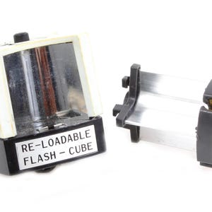 May include: Two vintage flash cubes, one assembled and one disassembled, against a white background. The assembled cube is black with a white label that reads "RE-LOADABLE FLASH - CUBE". The disassembled cube is silver and black.