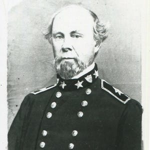 May include: A black and white portrait of a middle-aged individual with a receding hairline and a short, pointed beard. The subject wears a dark military-style uniform jacket featuring a high collar with three visible stars and metallic buttons.