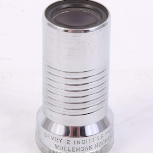 May include: A silver-colored Devry 2 inch F1.6 Wollensak Rochester lens. The lens has a cylindrical body with horizontal ridges and a wider base. The top of the lens is black.