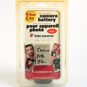 May include: A clear plastic package containing a rechargeable camera battery for a Canon NB 3L camera. The package is yellow and red with the text "2 Year/Ans exchange warranty" and "rechargeable camera battery". The package also includes the text "Pile rechargeable pour appareil photo NB5L" and "foto source".