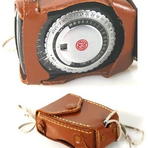 May include: A vintage General Electric light meter featuring a circular silver dial with black numerical markings, f-stop, and time scales, housed in a brown leather case with a snap closure and a thin, braided silver-coloured neck strap.