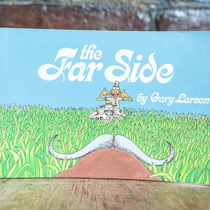 Op de afbeelding: Een rechthoekige boekomslag met een lichtblauwe achtergrond en de witte tekst The Far Side by Gary Larson. De illustratie toont een persoon met een safarihoed die een grote, grijze, gekrulde snor fotografeert die uit het groene gras steekt.