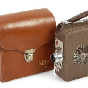 Puede incluir: Una funda de cámara de cuero marrón con un cierre plateado y el texto "DeJur 8" en la parte delantera. La funda está junto a una cámara de cine vintage de 8 mm marrón y plateada con el texto "Citation" en la parte delantera.