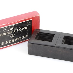 Puede incluir: Un adaptador de diapositivas 2x2 de plástico negro para un proyector. El adaptador está en una caja roja con el texto "20 BAUSCH & LOMB 2x2 ADAPTERS" impreso en él.