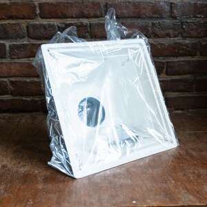 May include: A new, white, square sink with a circular drain hole, wrapped in clear plastic. The sink is on a wooden surface, with a brick wall in the background.