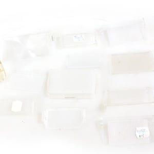 May include: A collection of clear plastic components, including rectangular and curved shapes, scattered on a white surface. Some pieces have small labels attached. A small, tan-colored plastic bracket is also present.