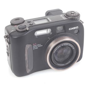 Może przedstawiać: Czarny aparat cyfrowy Casio z srebrnym pierścieniem obiektywu i wbudowaną lampą błyskową. Aparat ma rozdzielczość 4,1 megapiksela, widoczny jest numer modelu DV-4000. Na białym tle.