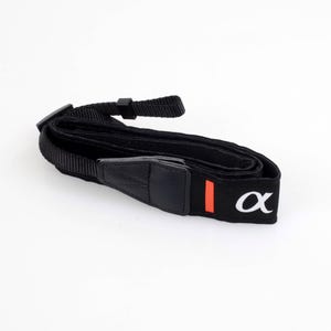 May include: Black camera strap with a white "α" logo and a red vertical stripe. The strap is made of a woven material and has adjustable buckles. The strap is designed for use with cameras.