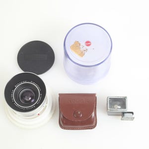 Pu&ograve; includere: Una collezione di attrezzature fotografiche vintage, tra cui un obiettivo, un tappo per l'obiettivo, una custodia in pelle marrone con la scritta "aquar" in rilievo, un accessorio flash e un contenitore di plastica trasparente. L'obiettivo ha dettagli neri e argentati.