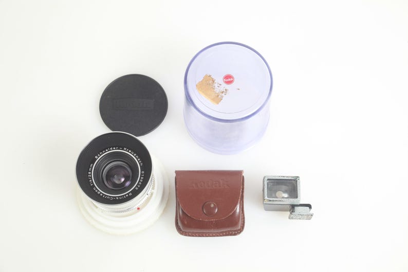 Pu&ograve; includere: Una collezione di attrezzature fotografiche vintage, tra cui un obiettivo, un tappo per l'obiettivo, una custodia in pelle marrone con la scritta "aquar" in rilievo, un accessorio flash e un contenitore di plastica trasparente. L'obiettivo ha dettagli neri e argentati.