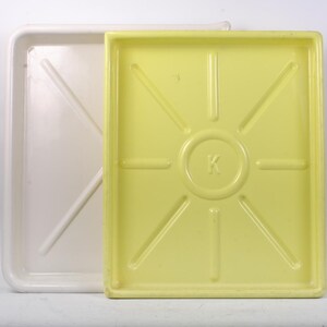 May include: Two rectangular trays. One is white with an X-shaped support in the center. The other is yellow with a sunburst design and the letter "K" in the center. Both trays have raised edges.