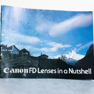 May include: A vintage Canon FD Lenses in a Nutshell booklet featuring a blue sky and mountain scene on the cover.