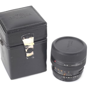 May include: Black camera lens with a protective cap and a black leather case. The lens has measurement markings and a red dot. The case has the brand name "Bronica" embossed on the top.