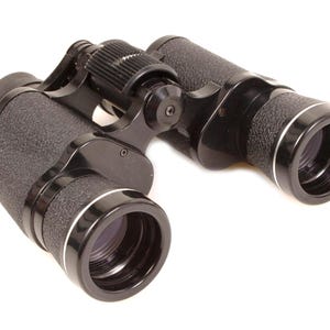 May include: Black binoculars with textured grips and silver trim around the lenses. The binoculars have a central focus wheel and are designed for viewing distant objects. The image is set against a white background.