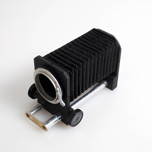May include: A black bellows camera attachment with a silver lens mount and support rods. The bellows are accordion-style, and the device sits on a white surface. The camera accessory is designed for macro photography.