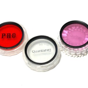Könnte beinhalten: Drei Objektivfilter für Kameras in durchsichtigen Plastikbehältern. Die Filter sind rot, rosa und klar. Der rote Filter hat den Schriftzug "PRO" aufgedruckt. Der klare Filter hat den Schriftzug "Quantaray" aufgedruckt.
