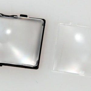 May include: Two clear, rectangular camera viewfinder accessories. One is in a black frame with mounting clips, the other is a separate clear lens. Both are on a white surface.