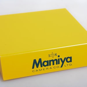 May include: A vibrant yellow display stand featuring the text "Mamiya CAMERA CO. LTD." in dark blue. The stand has a rectangular shape with a sloping front. The design is simple and contemporary.