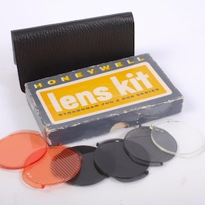 May include: A vintage Honeywell Lens Kit for Strobonar 700 & 800 series. The kit includes a black leather case, a box with the text "Lens Kit", and various colored and clear lens filters. The filters are orange, black, and clear.