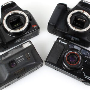 May include: Four black Canon cameras are displayed. Two are DSLR models, one marked "EOS", and the other two are point-and-shoot cameras. One point-and-shoot camera has "AUTO FOCUS" and "SELF-TIMER" printed on it.