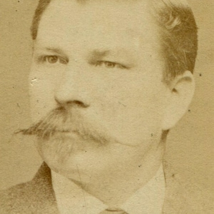 May include: Sepia-toned portrait of a man with a prominent mustache and neatly styled hair. He is wearing a suit with a collared shirt and a patterned tie. The photograph has a vintage aesthetic.