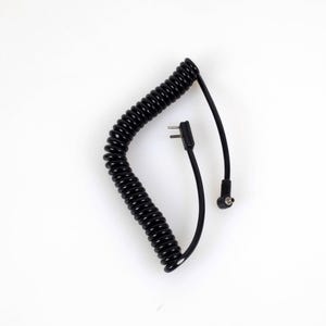 Puede incluir: Un cable negro en espiral con un diseño flexible similar a un resorte. El cable incluye dos extremos distintos: uno con un conector plano de dos clavijas y otro con un enchufe circular pequeño, ambos unidos a segmentos cortos de cable negro.