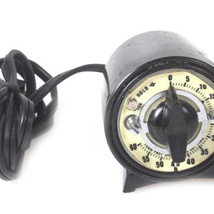 May include: A black cylindrical electric timer with a cream-colored circular dial featuring black numbers from 0 to 60. A black central knob controls the settings, with text reading HOLD, TRIGGER, and TURN PAST 0 THEN SET TIME.