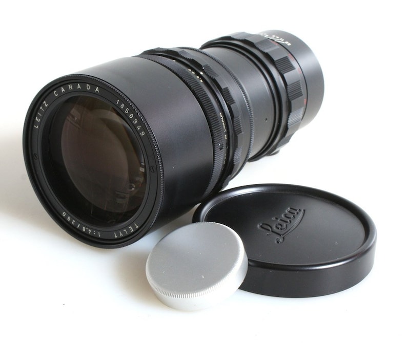 K&ouml;nnte beinhalten: Ein schwarzes Leica Tele-Elmarit 135mm f/3.5 Objektiv mit einer silbernen Objektivkappe und einer schwarzen Objektivkappe mit dem Leica Logo.