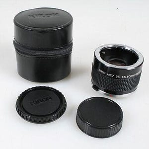 May include: A black Kiron MC7 2X teleconverter lens accessory with a textured grip, accompanied by a matching black Kiron branded lens cap, a secondary black lens cap, and a cylindrical black Kiron branded carrying case with a zipper closure.