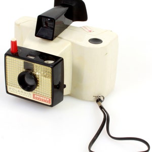 May include: A white and black Polaroid Swinger Model 85 Land Camera with a black leather strap. The camera has a red button on the side and a black viewfinder on top.
