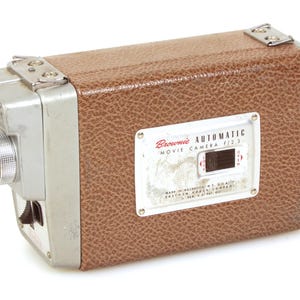 May include: Vintage Brownie Automatic Movie Camera f/2.3. The camera has a brown textured body with silver metal accents and a lens. The camera has a label that reads "Brownie Automatic Movie Camera f/2.3".
