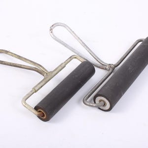 May include: Two vintage printmaking brayers. Each brayer has a dark grey roller and a silver-coloured metal handle. The rollers are cylindrical and appear to be made of a dense rubber-like material. The handles have a loop at the top.