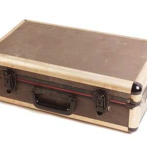 Puede incluir: Una maleta de transporte de metal marrón con un asa negra y dos cierres negros. La maleta tiene un ribete plateado y una franja roja.