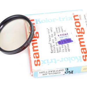 May include: A black Samigon circular diffraction filter with a handle, next to a vintage box. The box is labeled "Kolor-trix" and "Samigon" in red and orange, with text about the filter's quality.