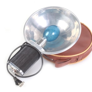 May include: Vintage photographic flash unit with a silver reflector, blue bulb, and black battery pack. Includes a brown leather carrying case with a zipper and handle. The unit is designed for photography.