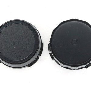 May include: Two black lens caps for a camera. One lens cap has a textured surface and the other has a smooth surface with a small circle in the center.