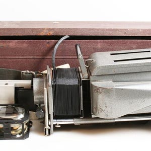 May include: Vintage silver-toned projector with a black bellows and a brown wooden box. The projector has a lens, a handle with a blue tip, and a green stripe. The box has a green strap.